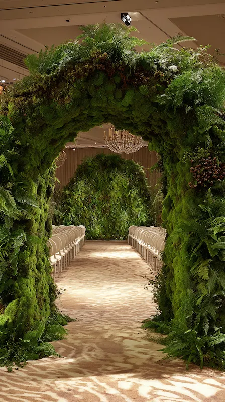 Luxury wedding ceremony aisle beneath towering moss and fern arch tunnel with white chairs leading to lush floral backdrop