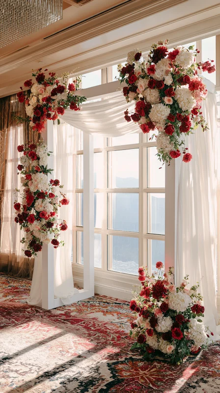 White wedding ceremony arch with cascading red and white flowers, soft draping, and natural light in elegant indoor venue
