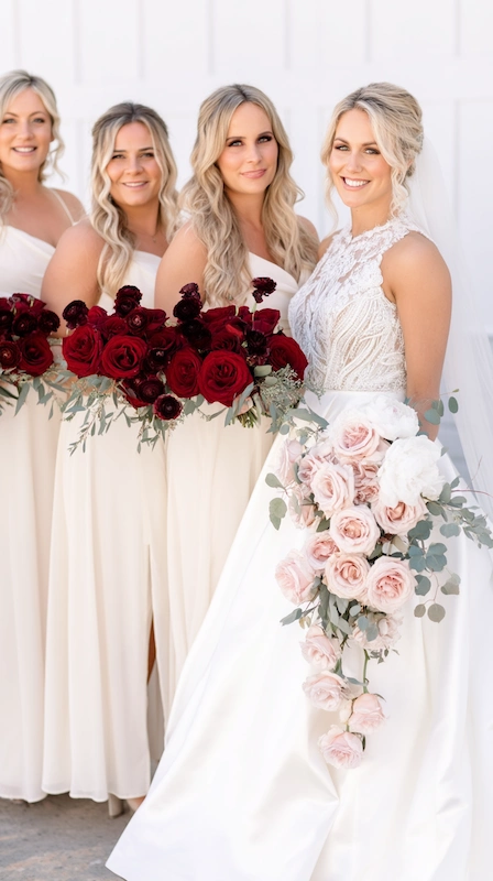 Bride in lace gown with cascading blush bouquet beside bridesmaids in neutral dresses holding deep red rose arrangements