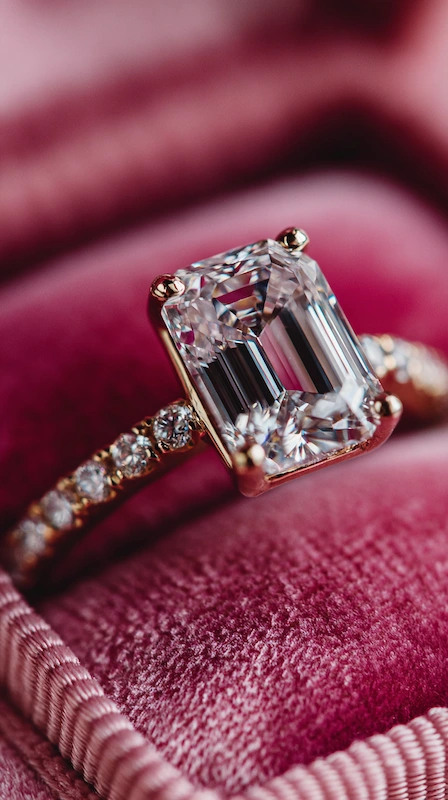 Emerald cut diamond engagement ring with pavé band resting on rich pink velvet with light reflecting through crisp facets
