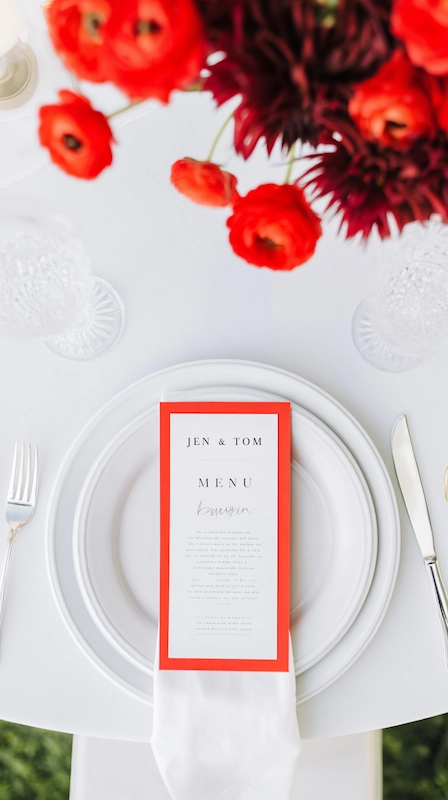 Modern wedding place setting with white plates, folded napkin, and menu card with red border, accented by bold red florals