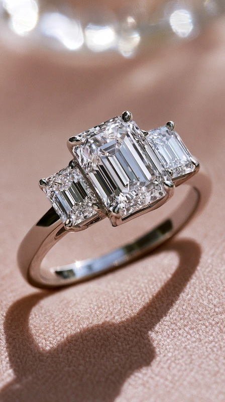 Popular Engagement Rings That Will Still Turn Heads in 20 Years 4 Emerald cut three stone diamond engagement ring on soft blush fabric, featuring trapezoid side stones and sleek polished band