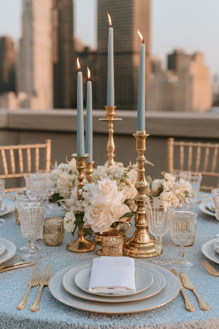 Dusty Blue Wedding Decor: Florals, Table Settings and Reception Styling 14 Dusty blue taper candles in brass candlesticks with white floral centerpiece on an elegant rooftop wedding reception table.
