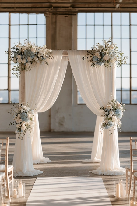 Dusty Blue Wedding Decor: Florals, Table Settings and Reception Styling 13 Dusty blue wedding ceremony backdrop with flowing ivory drapes, soft florals, candlelight, and a bright industrial venue.