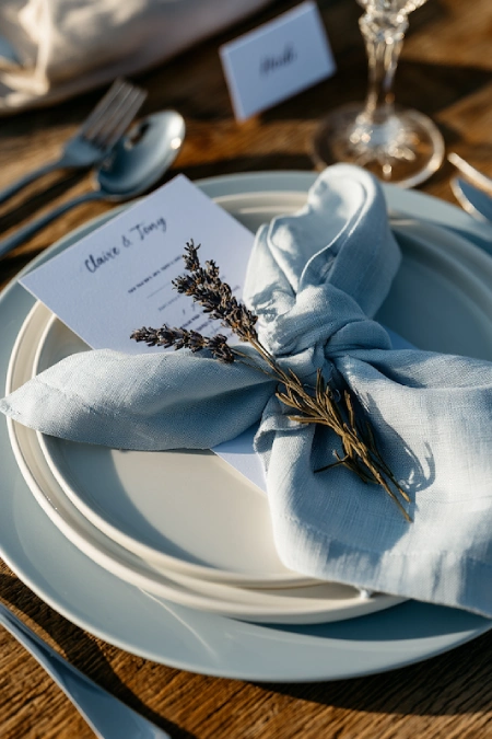 Dusty Blue Wedding Decor: Florals, Table Settings and Reception Styling 6 Dusty blue wedding place setting with layered white plates, linen napkin, lavender sprig, printed menu, and crystal stemware.