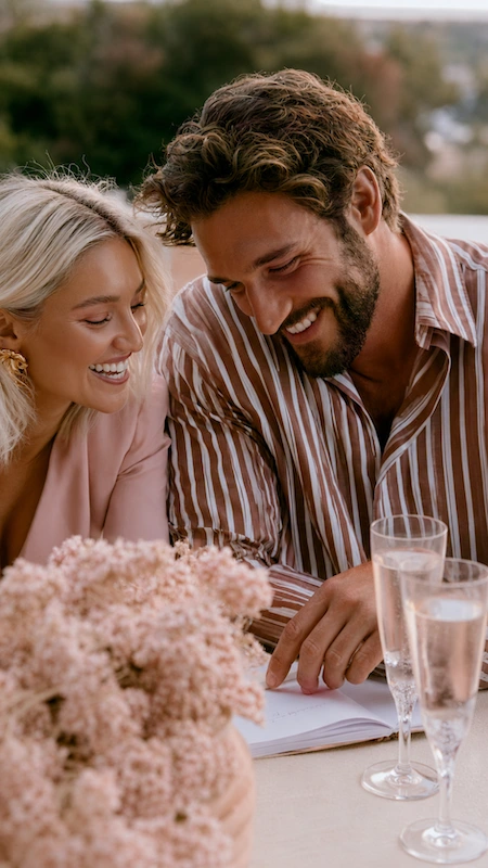 Newly engaged couple reviewing wedding planning notes with champagne and blush flowers, sharing a joyful moment together.