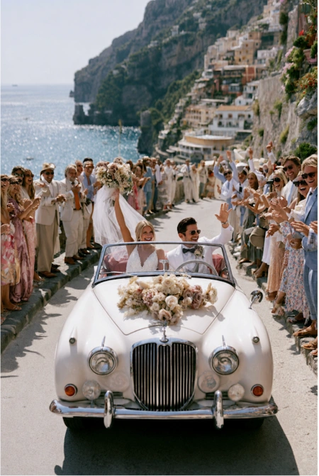 What to discuss before planning a wedding 6 Newlyweds ride through a coastal village in a vintage convertible as wedding guests cheer along the scenic waterfront
