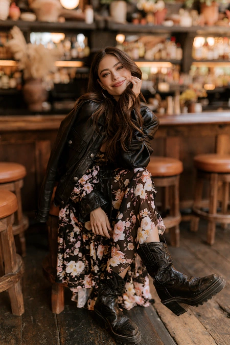 Woman in a floral maxi dress with a leather jacket and boots at a casual rehearsal dinner in a cozy restaurant setting