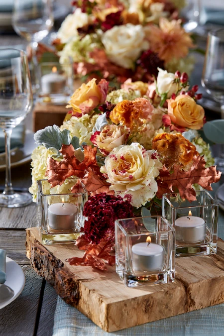 Fall wedding centerpiece with cream roses, rust florals, autumn leaves, candles, and wood risers on a rustic reception table