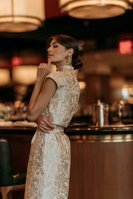 Bride-to-be in a gold brocade high neck dress with cap sleeves and sleek bun posing in an upscale rehearsal dinner restaurant