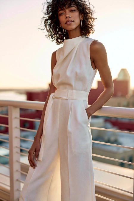 Bride-to-be in a sleek ivory jumpsuit with high neckline and belted waist posing on a rooftop terrace at golden hour