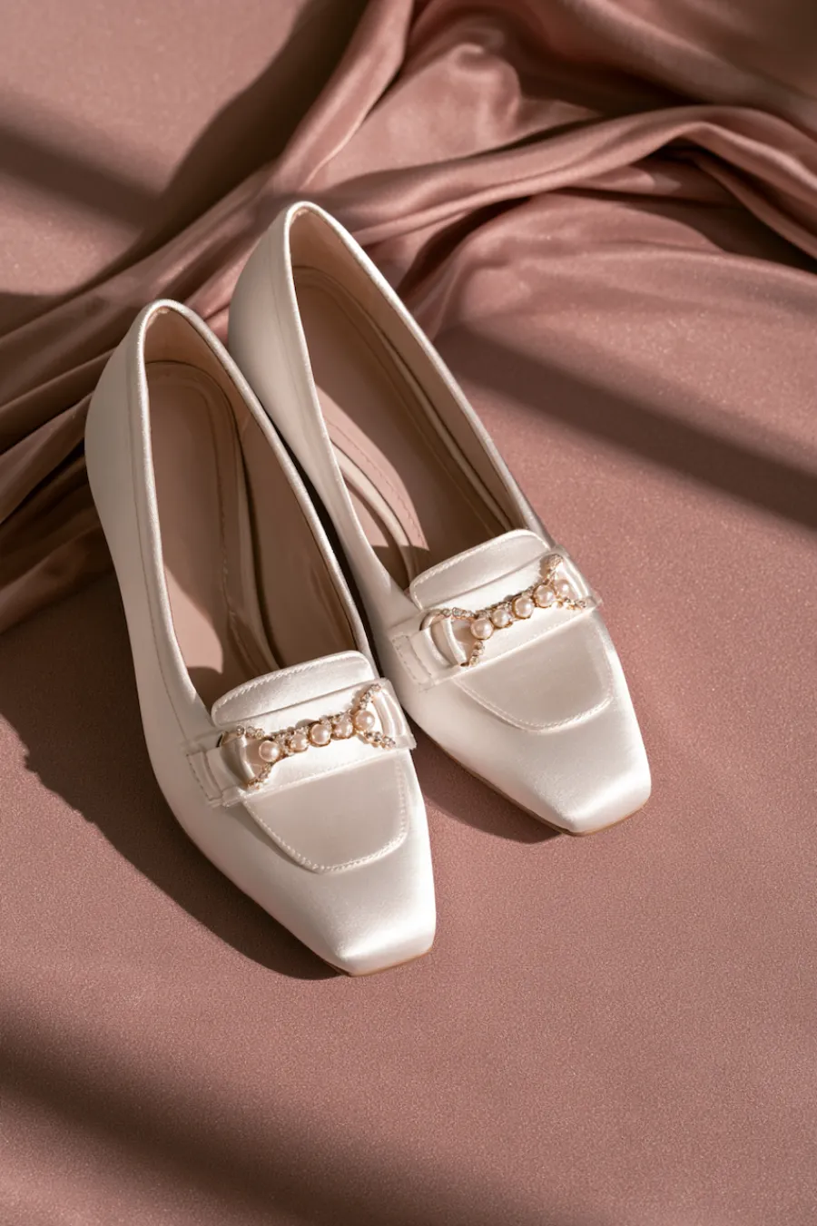 Wedding Shoes for the Bride: 27 Beautiful Ideas That Balance Style and Comfort 19 Ivory satin bridal flats with square toes and pearl chain embellishments displayed on blush fabric for elegant wedding style