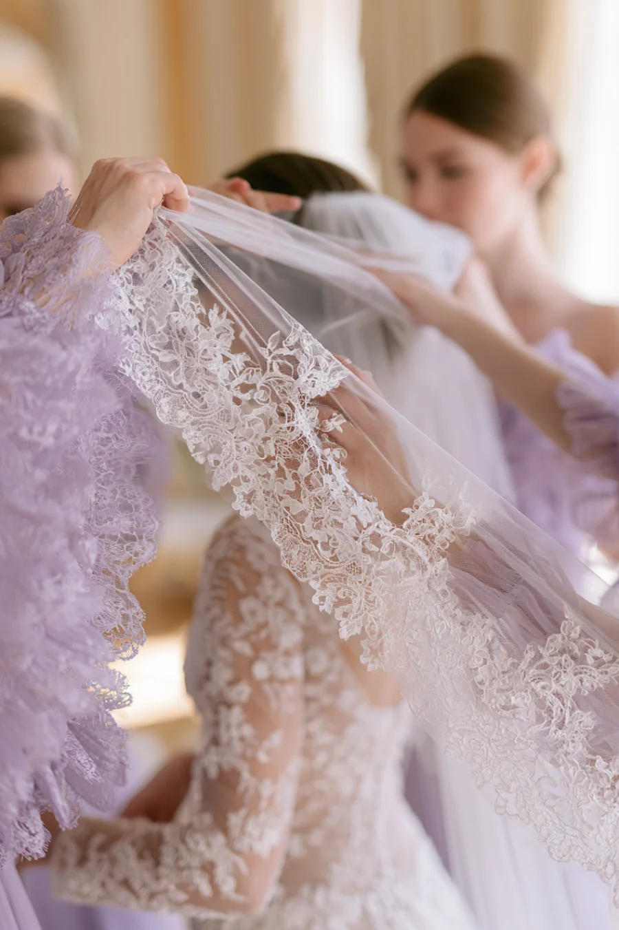 12 Timeless Lace Wedding Dress Styles We Love 17 Bride wearing a lace wedding dress as attendants place a lace-edged veil over her during elegant bridal wedding preparations