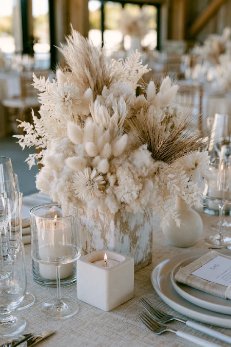Neutral dried floral centerpiece with pampas grass, bunny tails, and candles on an elegant rustic wedding reception table
