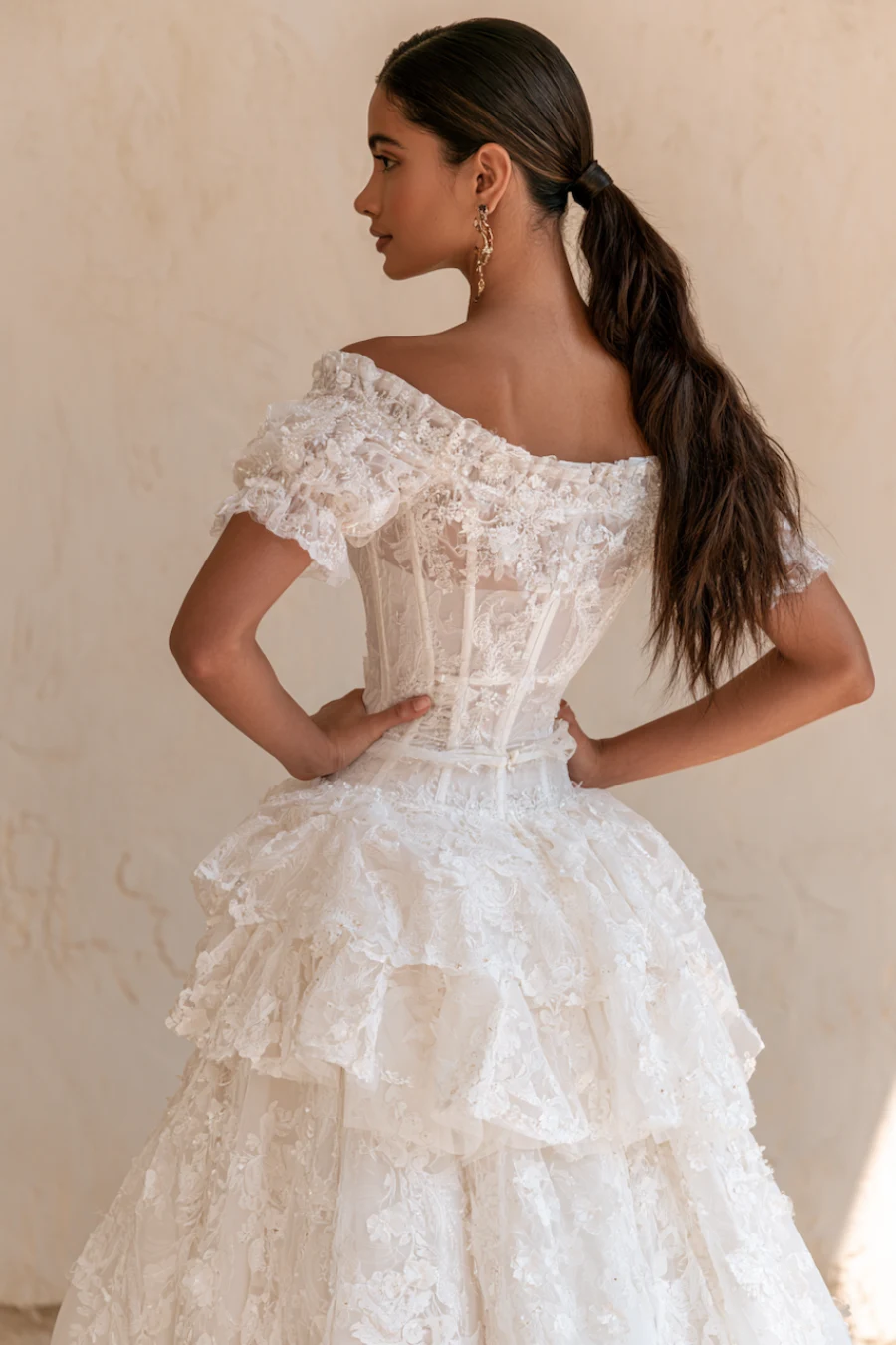 12 Timeless Lace Wedding Dress Styles We Love 10 Bride wearing an off-the-shoulder lace corset wedding dress with layered ruffles and floral appliqué viewed from the back