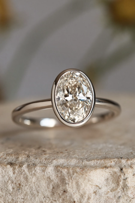 Oval Engagement Ring Obsession: Why This Cut Is Everyone's Main Character Right Now 13 Oval bezel set diamond engagement ring with a sleek white gold band displayed on natural stone in soft neutral light background