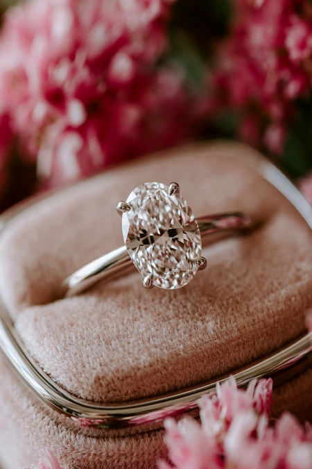 Oval Engagement Ring Obsession: Why This Cut Is Everyone's Main Character Right Now 9 Close-up of an oval diamond solitaire engagement ring on a blush velvet ring box with soft pink flowers in the background