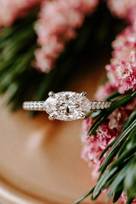 Oval Engagement Ring Obsession: Why This Cut Is Everyone's Main Character Right Now 11 Oval diamond solitaire engagement ring with pavé band nestled among soft pink flowers in warm natural light for a romantic look
