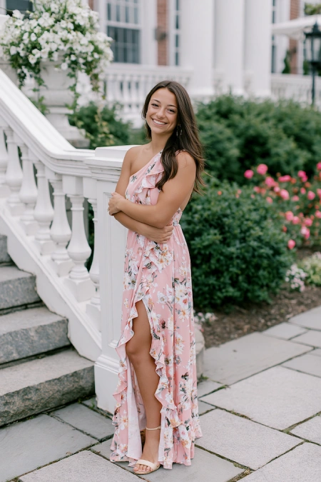 Bride-to-be in a pink floral one shoulder maxi dress with ruffle slit posing by a grand staircase before her rehearsal dinner