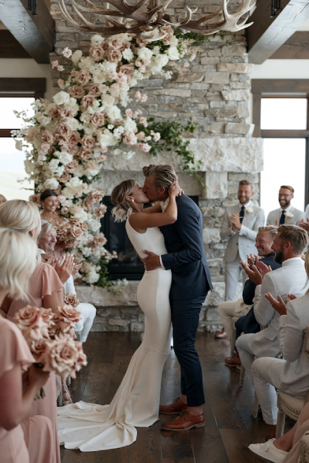 Sage Green and Terracotta Wedding: The Earthy Duo That Understood the Assignment 7 Bride and groom share their first kiss beneath a blush floral ceremony arch in a rustic stone venue surrounded by applauding guests