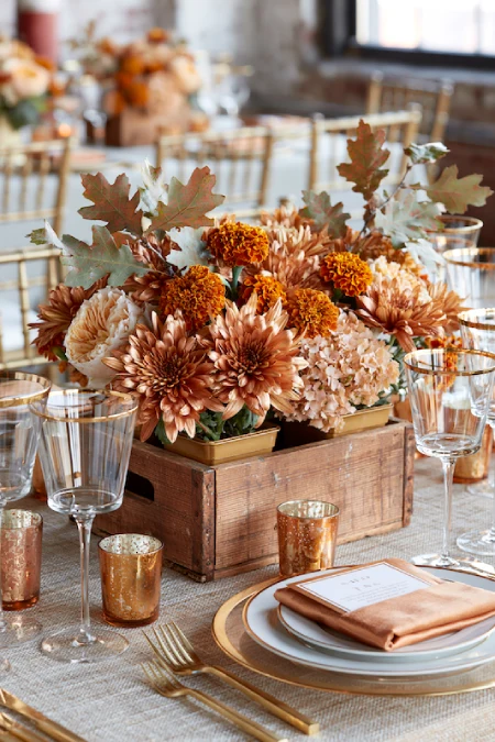 Rustic wooden box centerpiece with bronze mums, marigolds, hydrangeas, and autumn leaves on an elegant fall wedding table