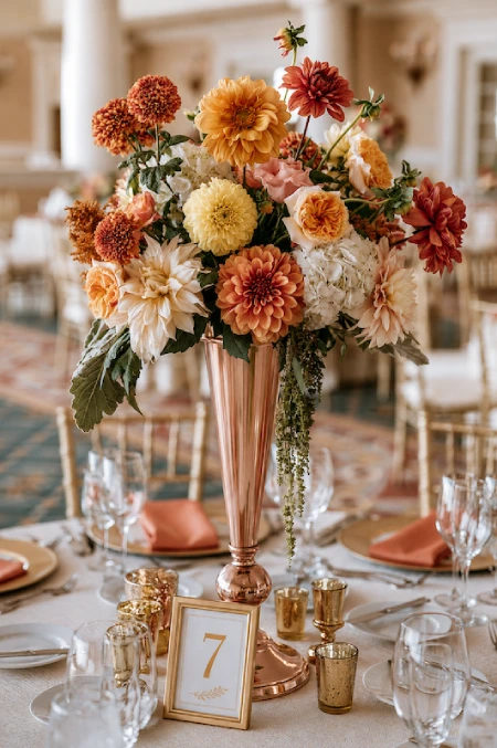 Tall copper vase centerpiece with caramel dahlias, roses, hydrangeas, and autumn blooms on an elegant wedding reception table