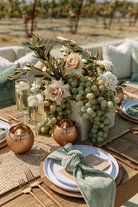 Vineyard wedding centerpiece with ivory roses, cascading green grapes, olive branches, and candlelight on an outdoor table