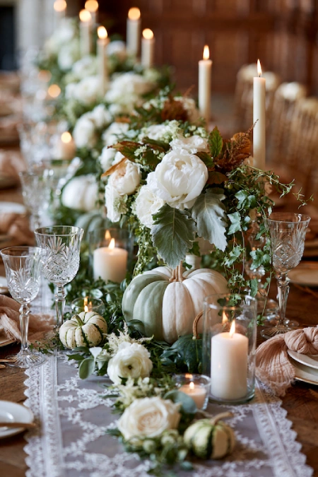 White pumpkin centerpiece with ivory roses, lush greenery, and pillar candles on an elegant fall wedding reception table