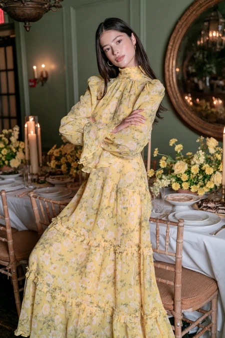 Bride-to-be in a yellow floral chiffon maxi dress with high neck and ruffles at a candlelit rehearsal dinner with yellow roses