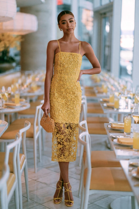 Bride-to-be in a yellow lace midi dress with sheer hem and matching heels standing in a bright modern rehearsal dinner venue