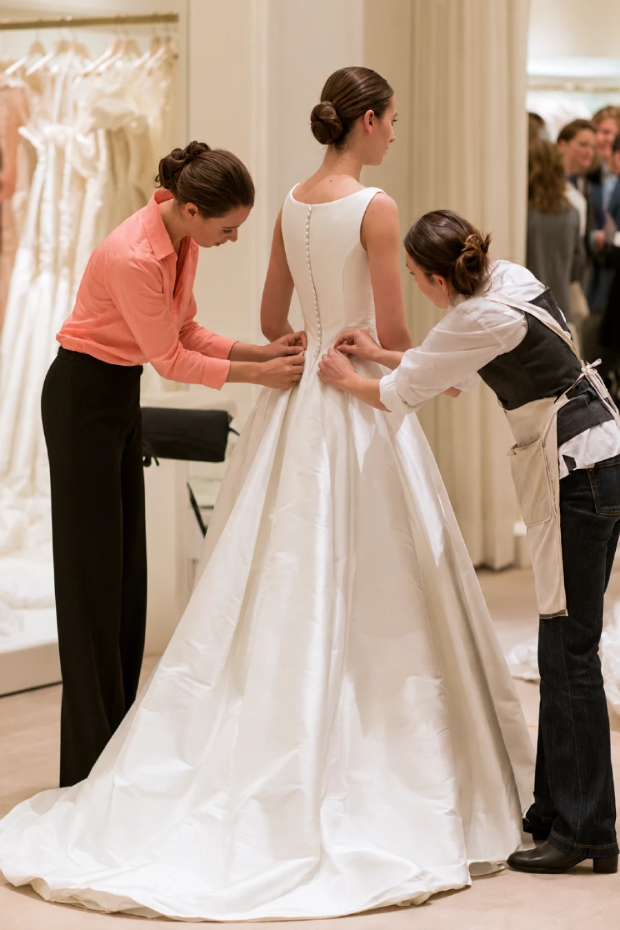 6-Month Wedding Planning Timeline - How to Plan a Wedding in 6 Months 4 Bride in a white satin wedding gown at her fitting, with two seamstresses adjusting the waist and bodice in a bridal boutique